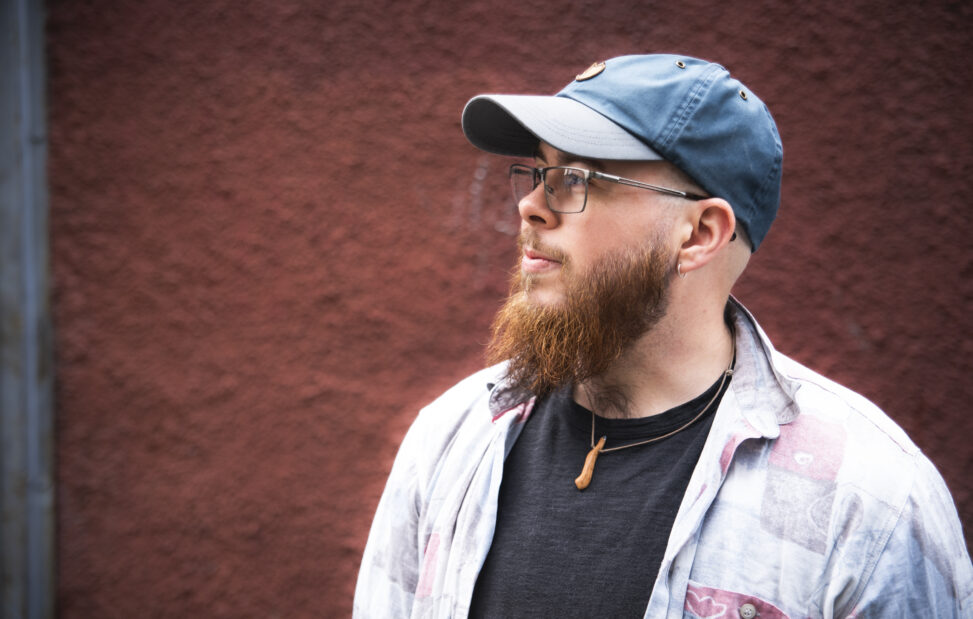 Max Alexander, a white man in his 30s with a long beard, he's wearing a dark blue baseball cap, shirt & T-shirt.