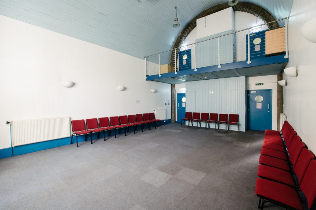 Photo showing the interior of a high community style hall. There are carpet tiles, a curved roof and doors at one end. There is a large window (not shown) at the other end. There is also a small balcony or mezzanine area with 2 doors. The room is lined with loose red chairs. 