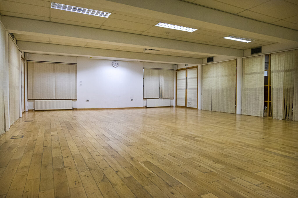 Photo of a low ceiling'd room with ceiling tiles and strip lights. There are windows and glass doors in the space with blinds covering each. 